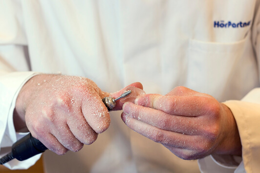 Ein Hörakustiker bearbeitet ein Hörgerät-Handstück präzise mit einem Fräswerkzeug.